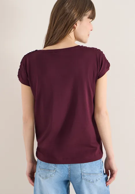 Frau steht mit dem Rücken zur Kamera, trägt ein kastanienbraunes T-Shirt und hellblaue Jeans.